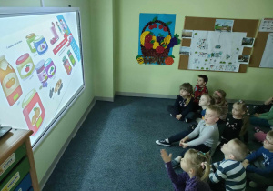 Dzieci z grupy "Pszczółek" oglądają film edukacyjny pt. "Skąd się biorą produkty ekologiczne?" na tablicy interaktywnej.
