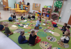 "Czerwony Kapturek" w "Pszczółkach"-dzieci siedzą na dywanie i odgadują zagadkę o jabłku. Chłopiec podnosi jabłko. Pozostałe dzieci obserwują.