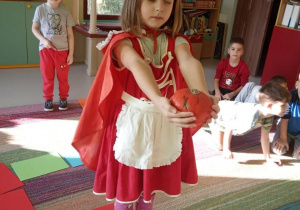"Czerwony Kapturek" w "Kaczuszkach"-dziewczynka w stroju czerwonego kapturka z dynią w ręku stoi na białym polu i ma do wykonania zadanie.