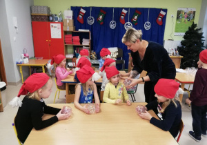 Dzieci z grupy "Kaczuszek" przy stołach słuchają instrukcji, w jaki sposób ozdobić bombki.