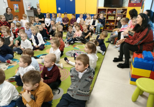 Przedszkolaki siedzą na dywanie i słuchają muzyki, pochodzącej z instrumentu.