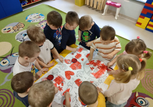 Dzieci z grupy Pszczółek na dywanie podczas wspólnej pracy plastycznej, polegającej na malowaniu patyczkiem higienicznym kolorowanki w dużym formacie, przy użyciu czerwonej farby.