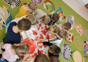 Dzieci z grupy Pszczółek na dywanie podczas wspólnej pracy plastycznej, polegającej na malowaniu patyczkiem higienicznym kolorowanki w dużym formacie, przy użyciu czerwonej farby.