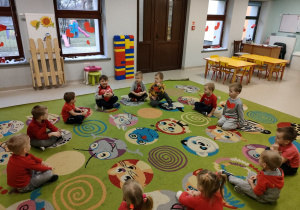 Dzieci z grupy Pszczółek siedzą w kole na dywanie w trakcie zabawy "Lubie Cię za to, że..." z wykorzystaniem czerwonego balona w kształcie serca.