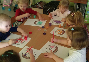 Kilkoro dzieci z grupy "Motylków" przy stoliku maluje szablon pączka za pomocą pędzelków i farb w różnych kolorach.