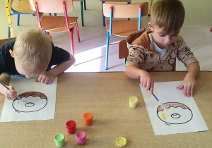 Dwóch chłopców z grupy "Motylków" przy stoliku maluje szablon pączka za pomocą pędzelków i farb w różnych kolorach.