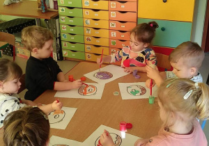 Kilkoro dzieci z grupy "Motylków" przy stoliku maluje szablon pączka za pomocą pędzelków i farb w różnych kolorach.