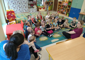Pani Ola opowiada dzieciom o swoim zawodzie i miejscu, w jakim pracuje. Dzieci siedzą na dywanie i uważnie słuchają.