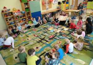 Pani bibliotekarka prezentuje dzieciom różne rodzaje książek. Dzieci siedzą w kole na dywanie i uważnie oglądają.