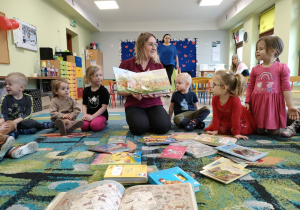 Pani bibliotekarka prezentuje dzieciom różne rodzaje książek. Dzieci siedzą w kole na dywanie i uważnie oglądają.