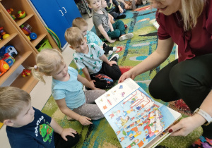 Pani Ola pokazuje dzieciom książkę. Zadaniem kilku dzieci jest wyszukanie danego elementu na obrazku.