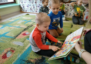 Pani Ola pokazuje dzieciom książkę. Zadaniem kilku dzieci jest wyszukanie danego elementu na obrazku.