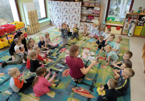 Pani Aleksandra prezentuje dzieciom różne książeczki, w których ukryte zostały zadania. Dzieci siedzą w kole i uważnie słuchają oraz oglądają.