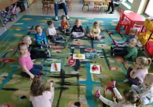 Dzieci z grupy "Motylków" siedzą na dywanie w trakcie dopasowywania jabłka, marchewki i cytryny do ilustracji.