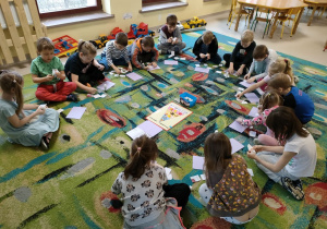 Dzieci z grupy "Skrzatów" siedzą na dywanie w trakcie naklejania złożonych obrazków warzyw/owoców na kartkę w jak najszybszym tempie.