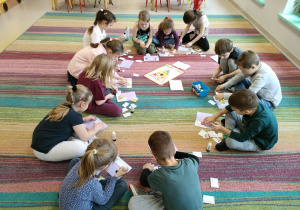 Dzieci z grupy "Kaczuszek" siedzą na dywanie w trakcie naklejania złożonych obrazków warzyw/owoców na kartkę w jak najszybszym tempie.