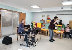 Jeden prowadzący gra na instrumencie, natomiast drugi prowadzący pokazuje dzieciom, jak dyrygować.