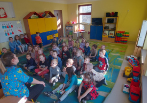 Dzieci z grupy ''Skrzaty'', ''Kaczuszki'' i ''Biedronki'' siedząc na dywanie z uwagą słuchają pani Aleksandry oraz jej asystentki.