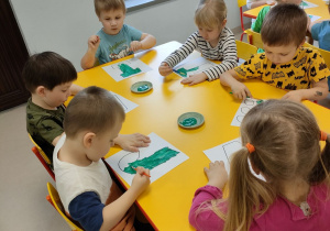 Kilkoro dzieci przy stole maluje szablon liter przy pomocy zielonej farby i pędzelków.
