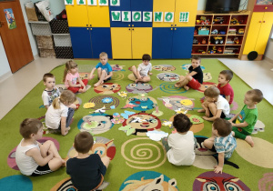 Dzieci siedzą na dywanie podczas pogadanki o symbolach wiosny oczekując na gimnastykę.