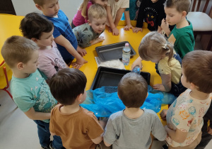 Dzieci z grupy "Pszczółek" w trakcie eksperymentu, polegającego na obserwacji wchłaniania wody przez różne materiały. Roksana polewa wodą płaszcz przeciwdeszczowy.