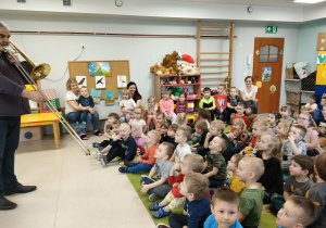 Pan Sławek prezentuje dzieciom grę na puzonie. Dzieci z uwagą oglądają i słuchają.
