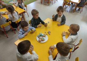 Kilkoro dzieci z grupy "Pszczółek" przy stoliku podczas poczęstunku w ramach wielkanocnego spotkania z Księdzem. Dzieci otrzymały przygotowane i udekorowane przez siebie babeczki oraz słodkie, czekoladowe jajka.