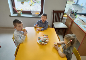 Kilkoro dzieci z grupy "Pszczółek" przy stoliku podczas poczęstunku w ramach wielkanocnego spotkania z Księdzem. Dzieci otrzymały przygotowane i udekorowane przez siebie babeczki oraz słodkie, czekoladowe jajka.