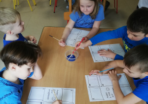 Grupa dzieci siedzi przy stolikach i maluje niebieską farbą.