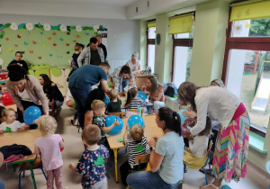 Dzieci wraz z Rodzicami ozdabiają kolorowe baloniki przy stołach.