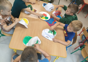 Kilkoro dzieci z grup młodszych przy stoliku stempelkuje/maluje czapkę przy pomocy gąbeczek i farb w różnych kolorach.