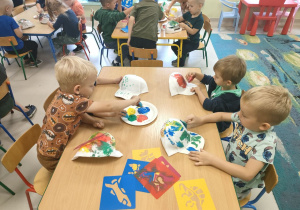 Kilkoro dzieci z grup młodszych przy stoliku stempelkuje/maluje czapkę przy pomocy gąbeczek i farb w różnych kolorach.