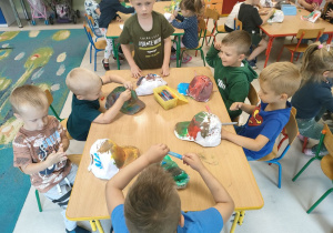Kilkoro dzieci z grup młodszych przy stoliku ozdabia czapkę za pomocą brokatowego kleju oraz kolorowych mazaków.