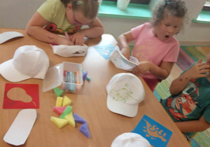Kilkoro dzieci z grup starszych ozdabia czapkę za pomocą brokatowego kleju oraz kolorowych mazaków.