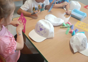 Kilkoro dzieci z grup starszych ozdabia czapkę za pomocą brokatowego kleju oraz kolorowych mazaków, korzystając także z różnych szablonów.