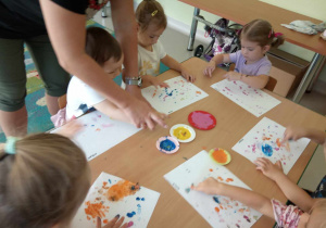 Kilkoro maluszków przy stole maluje kropki przy wykorzystaniu swoich paluszków i kolorowej farby.