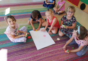 Kilkoro dzieci z grupy "Skrzatów" nakleja wycięte, kolorowe kółka na duży arkusz papieru.
