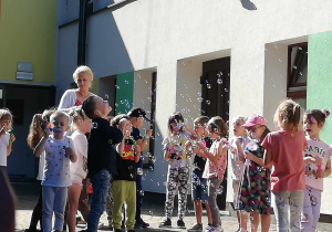 Ciocia Magda wraz z dziećmi z grupy "Skrzatów" puszcza bańki mydlane na tarasie przedszkolnym.