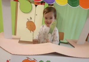 Dziewczynka pozuje do zdjęcia trzymając sylwetę warzyw na tle straganu warzywnego.