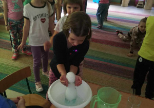 Dziewczynka z grupy "Skrzatów" podczas zabawy z pianką, utworzoną w wyniku połączenia ciepłej wody, kostek suchego lodu i mydła w płynie. Pozostałe dzieci czekają na swoją kolej.