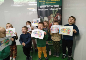 Kilkoro dzieci z grupy "Pszczółek" podczas sesji zdjęciowej ze swoimi pracami na tle baneru Hurtowni Warzyw i Owoców.