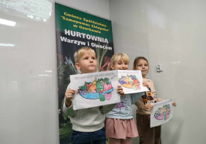 Artur, Klaudia i Zuzia z grupy "Pszczółek" podczas sesji zdjęciowej ze swoimi pracami na tle baneru Hurtowni Warzyw i Owoców.