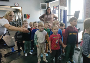 Pani Iwona Karczewska przedstawia dzieciom sprzęty na siłowni, tj. bieżnię i orbitrek.