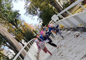 Dzieci z grupy "Pszczółek" biegną przez most nad rzeką "Bzurą" w parku miejskim.