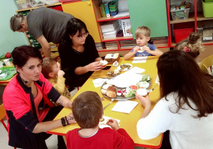 Dzieci siedzą przy stoliku wraz z Rodzicami podczas degustacji pysznych, domowych bułeczek z różnymi zdrowymi dodatkami.