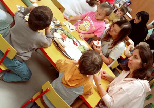 Dzieci siedzą przy stolikach wraz z Rodzicami podczas degustacji pysznych, domowych bułeczek z różnymi zdrowymi dodatkami.