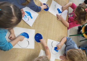 Kilkoro dzieci z grupy najmłodszej przy stoliku maluje szablon serca za pomocą paluszków i niebieskiej farby.