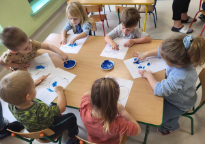 Kilkoro dzieci z grupy najmłodszej przy stoliku maluje szablon serca za pomocą paluszków i niebieskiej farby.