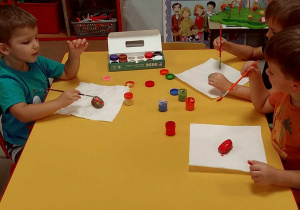 Kilkoro dzieci przy stoliku maluje kamienie za pomocą pędzelków i farby.