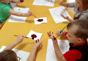 Kilkoro dzieci przy stole maluje dłonie pędzelkiem przy użyciu brązowej farby.